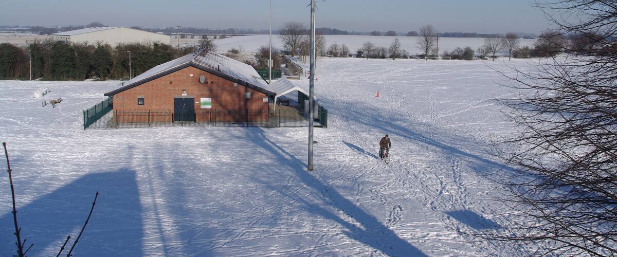 A snow-covered landscape with a brick building, a tall streetlight, and a person on a bicycle.