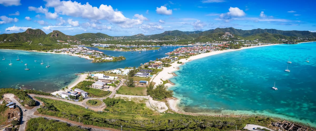 An aerial view of a coastal town with a beach, clear turquoise waters, and mountains in the background.