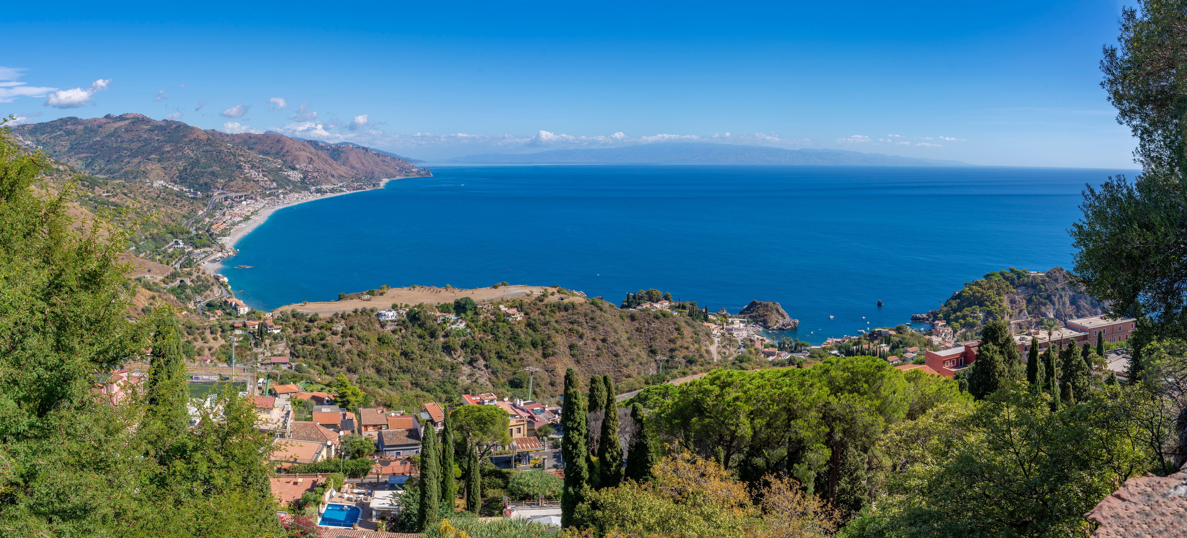 A coastal town with a clear view of the sea, surrounded by hills and dense greenery.