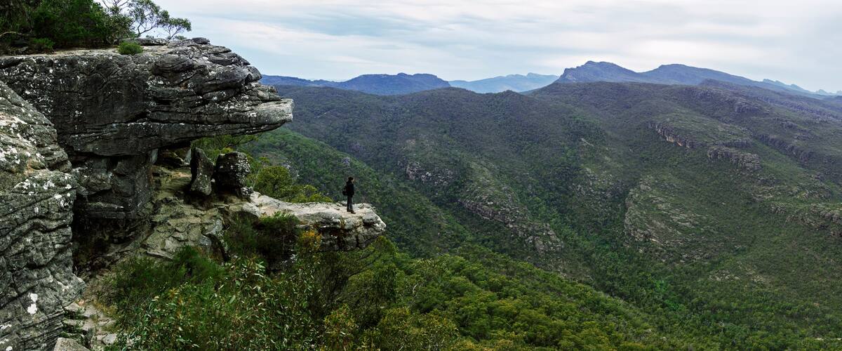 A mountainous landscape with a prominent rock formation on the left and a person standing on a cliff edge.