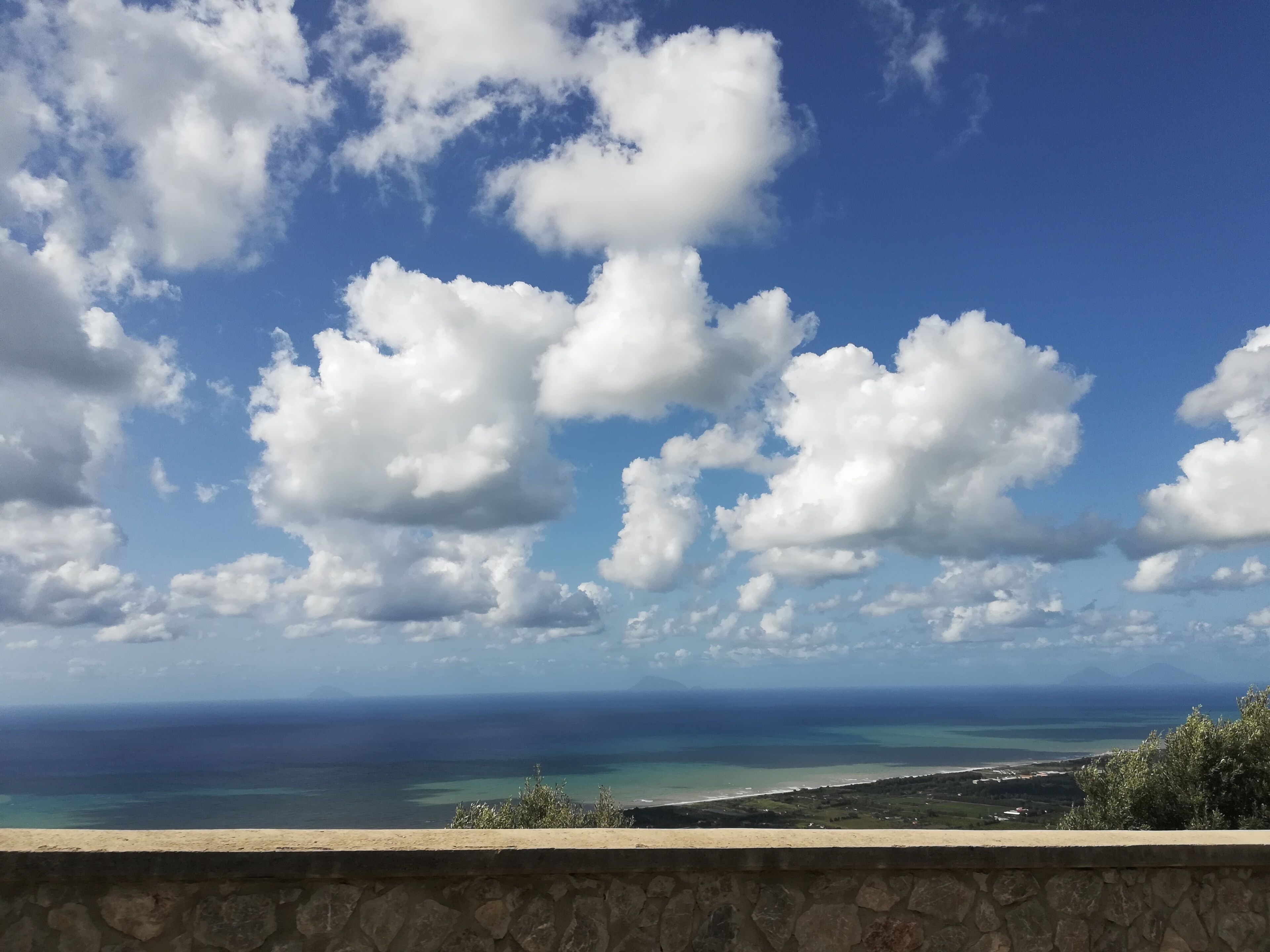 A coastal landscape with a stone wall in the foreground, a clear blue sky with scattered clouds, and a view of the ocean and distant land.