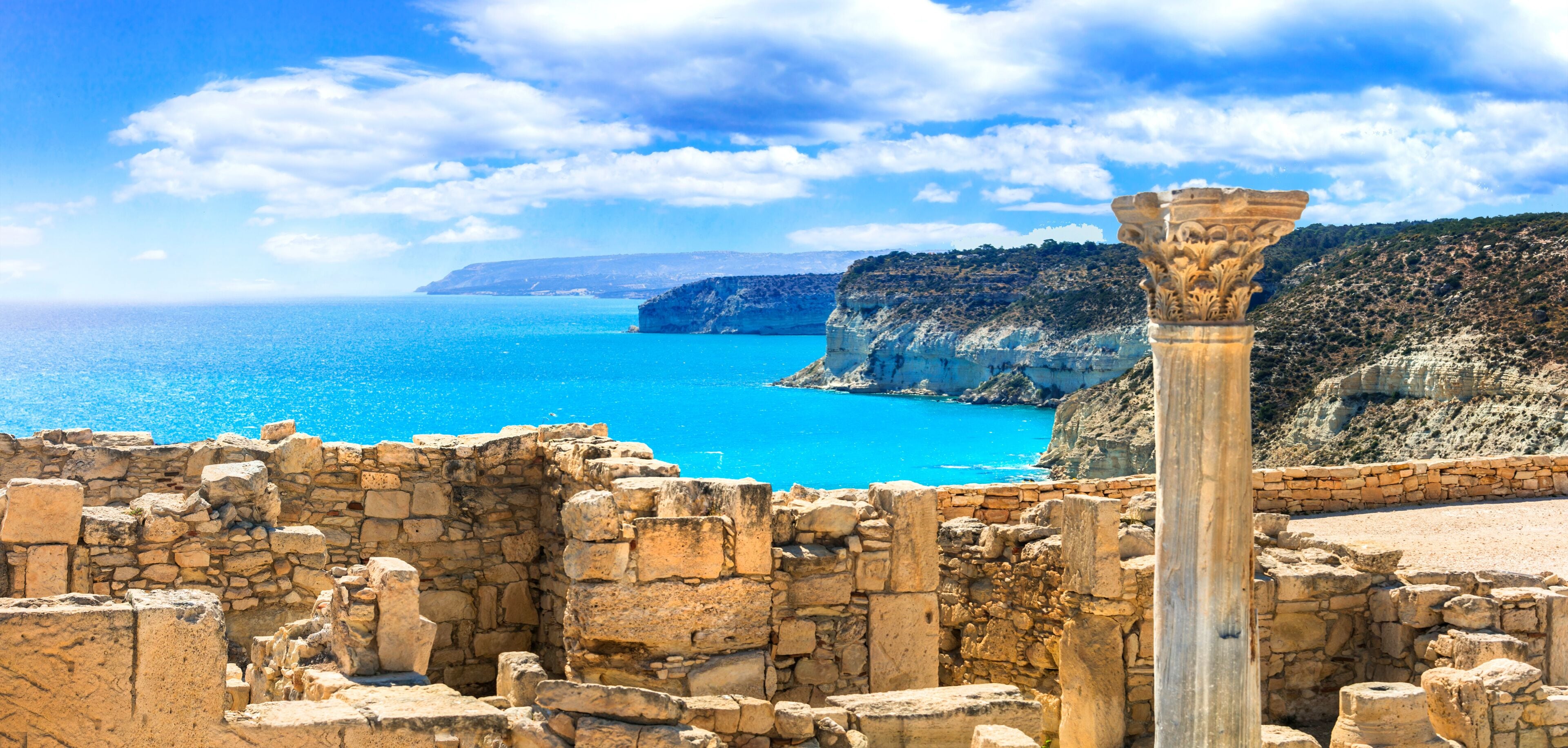Ancient ruins with a stone column, a clear blue sea, and a mountainous coastline.