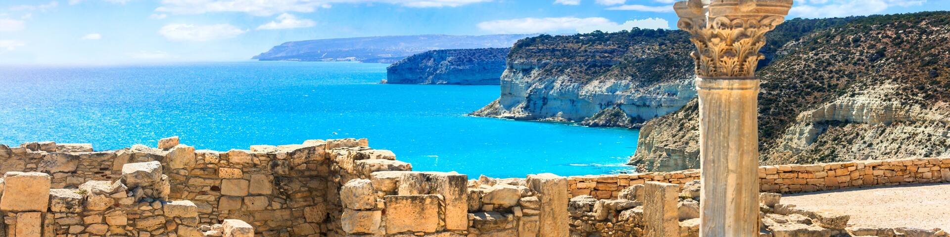 Ancient ruins with a stone column, a clear blue sea, and a mountainous coastline.