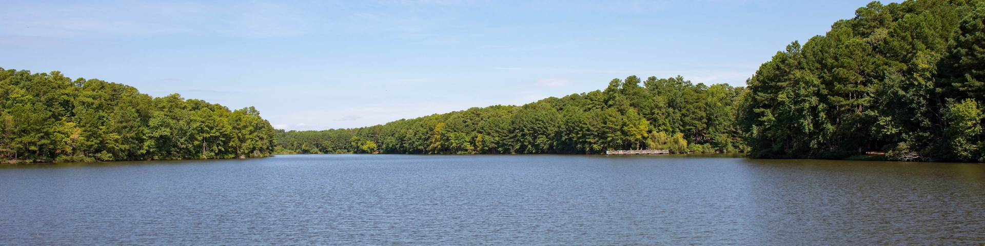A beautiful view of a lake with bright green trees and bright blue sky and a dock on the opposite shoreline.