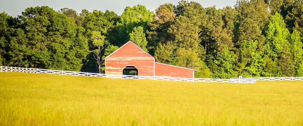 A red barn with a white fence in a grassy field, surrounded by dense green trees.