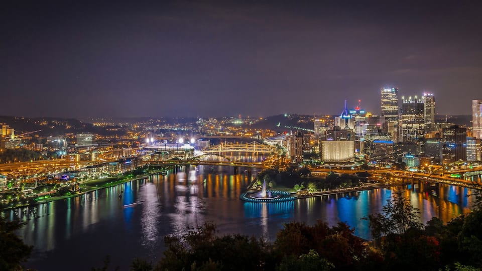 A nighttime cityscape with illuminated buildings, a river, and a bridge.