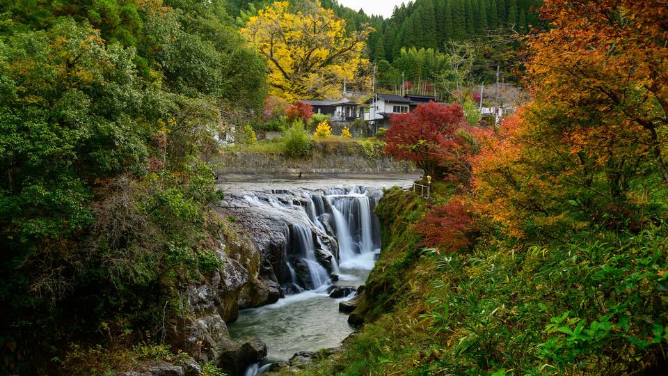 A waterfall surrounded by autumn foliage, with houses in the background.