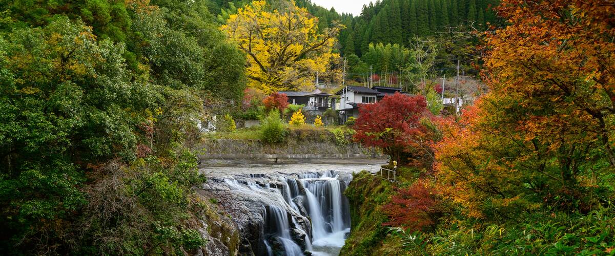 A waterfall surrounded by autumn foliage, with houses in the background.