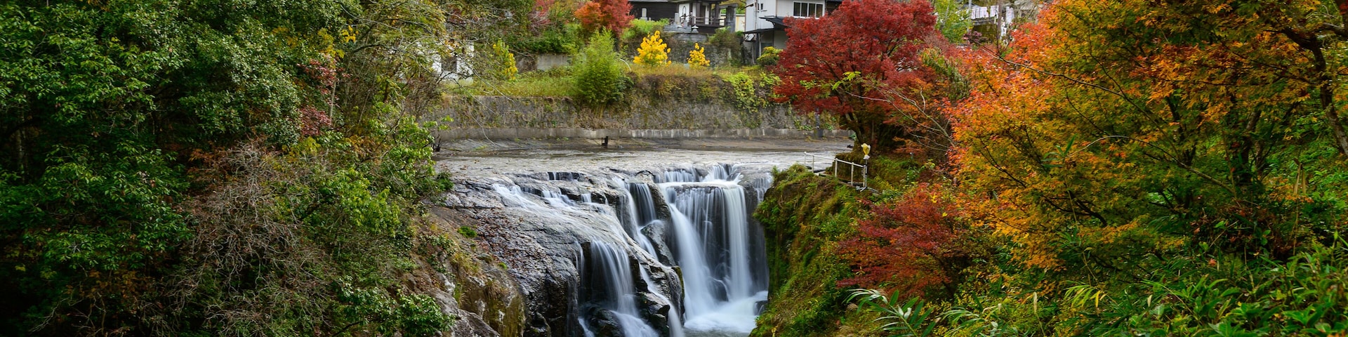 A waterfall surrounded by autumn foliage, with houses in the background.