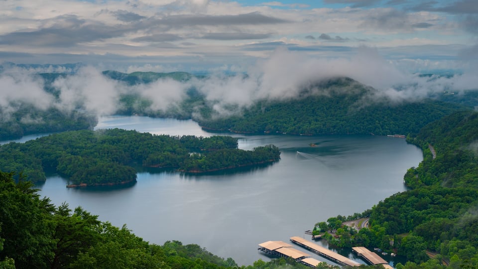 A lake surrounded by forested hills, with a small dock and buildings near the water's edge.
