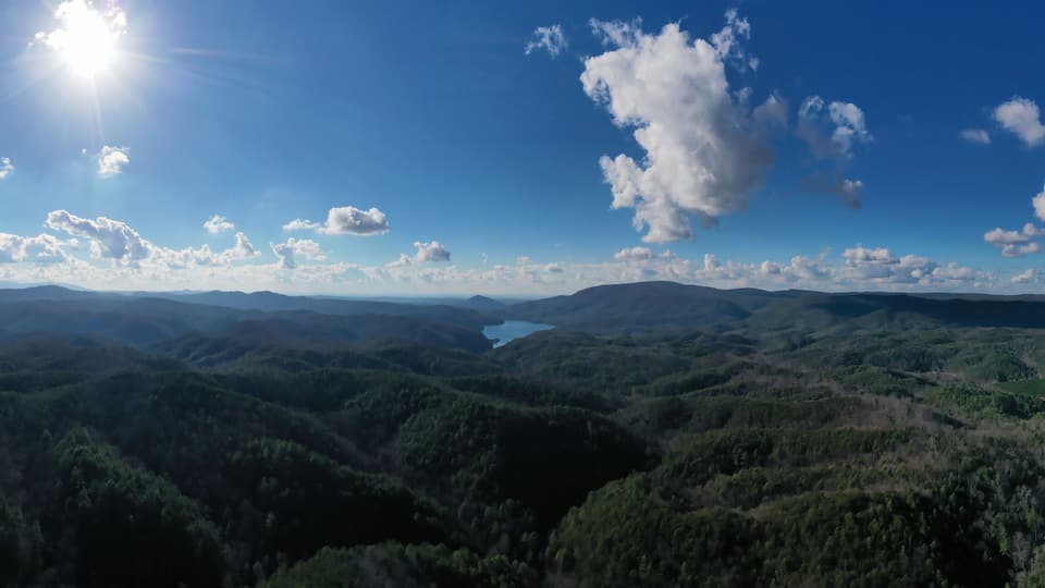A mountainous landscape with dense forests and a clear blue sky with scattered clouds.