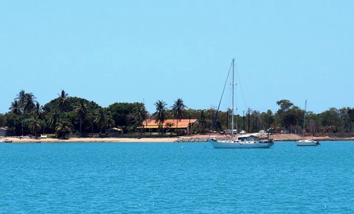 A coastal scene with a clear blue sea, a sailboat, and a beach with palm trees and buildings.