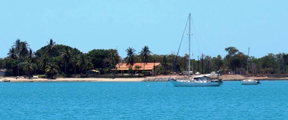 A coastal scene with a clear blue sea, a sailboat, and a beach with palm trees and buildings.