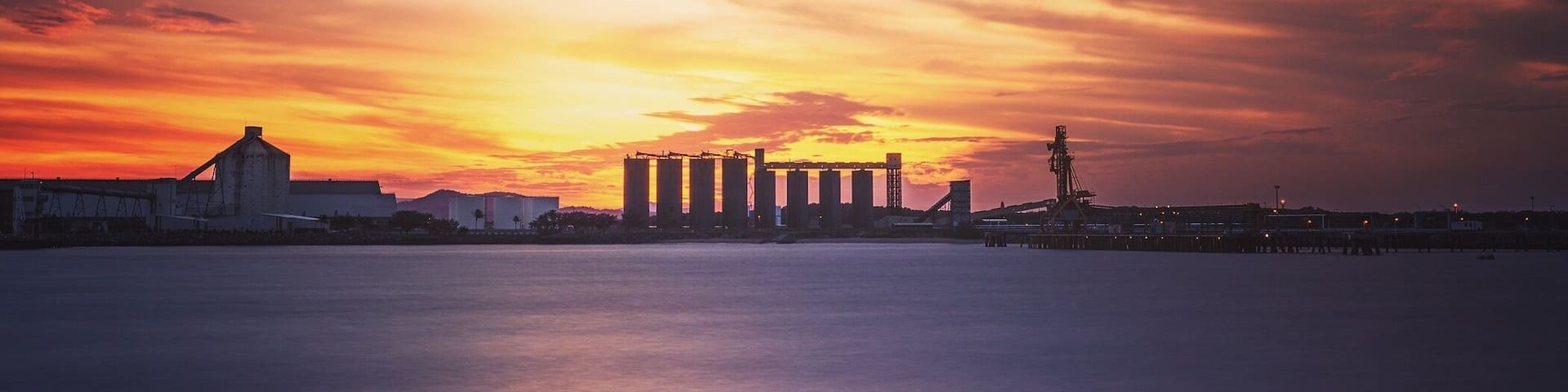 A sunset over a coastal industrial area with silos and cranes.