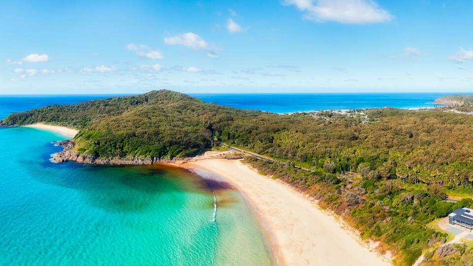 A coastal landscape with a beach, clear turquoise waters, and a hill with dense vegetation.