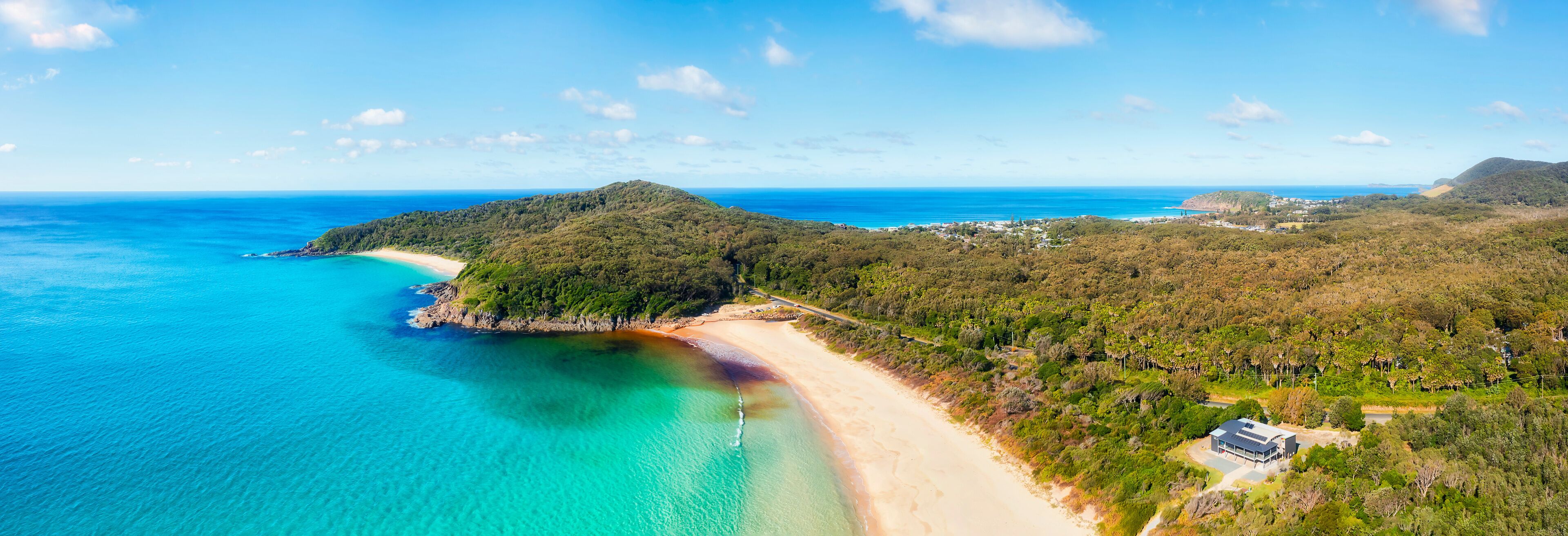 A coastal landscape with a beach, clear turquoise waters, and a hill with dense vegetation.