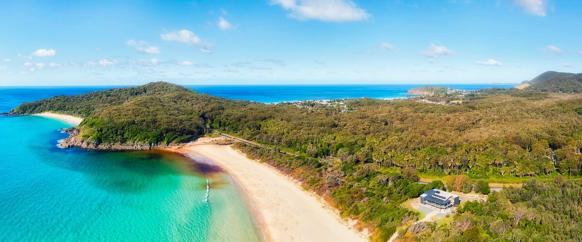 A coastal landscape with a beach, clear turquoise waters, and a hill with dense vegetation.