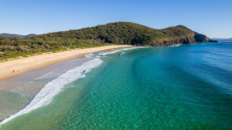 A beach with clear turquoise waters, a sandy shore, and a forested hill in the background.