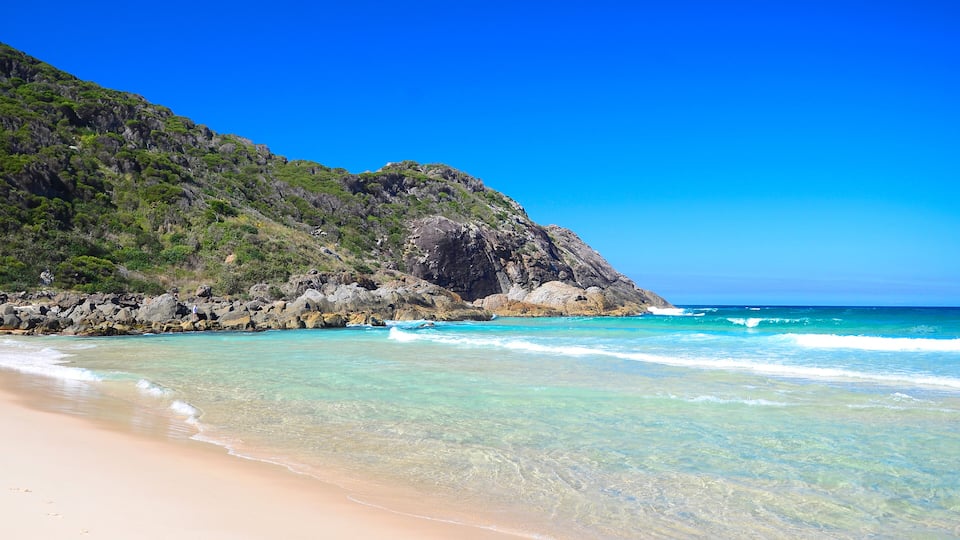 A beach with clear turquoise water, a sandy shore, and a rocky cliff with greenery.