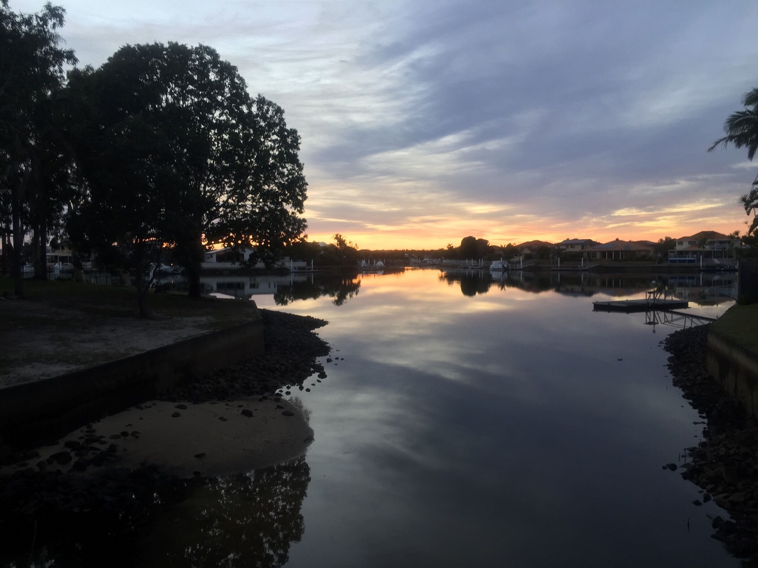 A calm lake at sunset with reflections of trees and buildings.