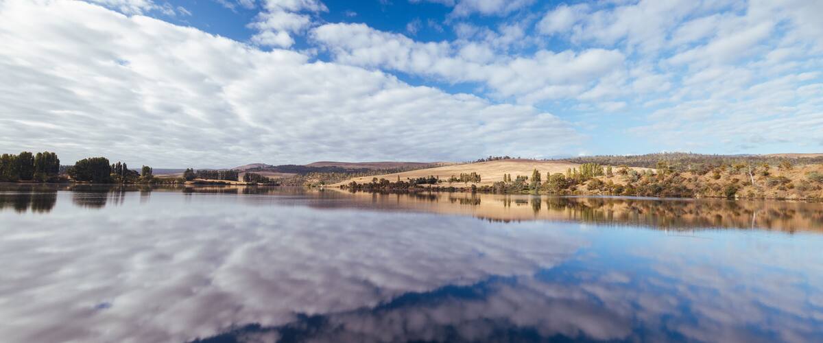 A calm lake reflecting clouds, surrounded by hills and sparse trees.