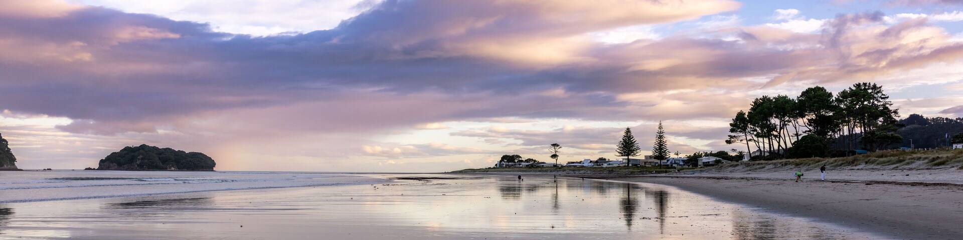 Beachgoers enjoy a tranquil evening walk on a reflective sandy Whangamata shore, with Clark Island and dramatic clouds mirrored in the wet sand. Coromandel Peninsula, New Zealand.