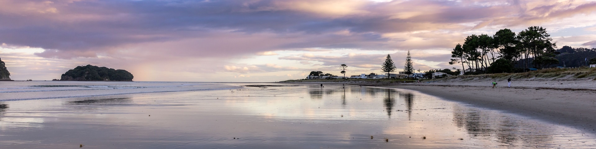 Beachgoers enjoy a tranquil evening walk on a reflective sandy Whangamata shore, with Clark Island and dramatic clouds mirrored in the wet sand. Coromandel Peninsula, New Zealand.
