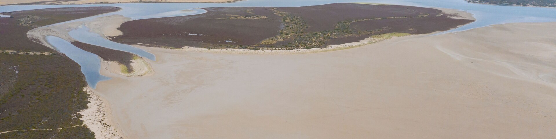 Un paisaje costero con una amplia zona arenosa, un canal de agua sinuoso y una costa distante con vegetación.