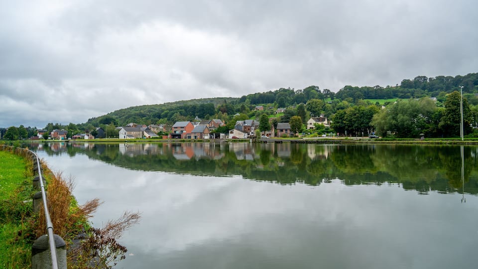 A calm lake with a reflection of houses and trees, surrounded by greenery and a cloudy sky.