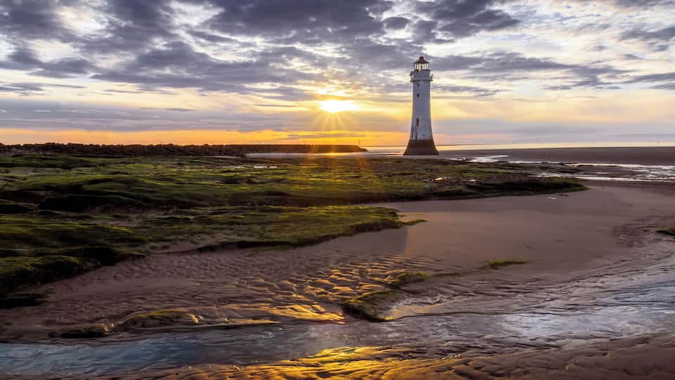 A lighthouse by a beach with a sunset in the background.
