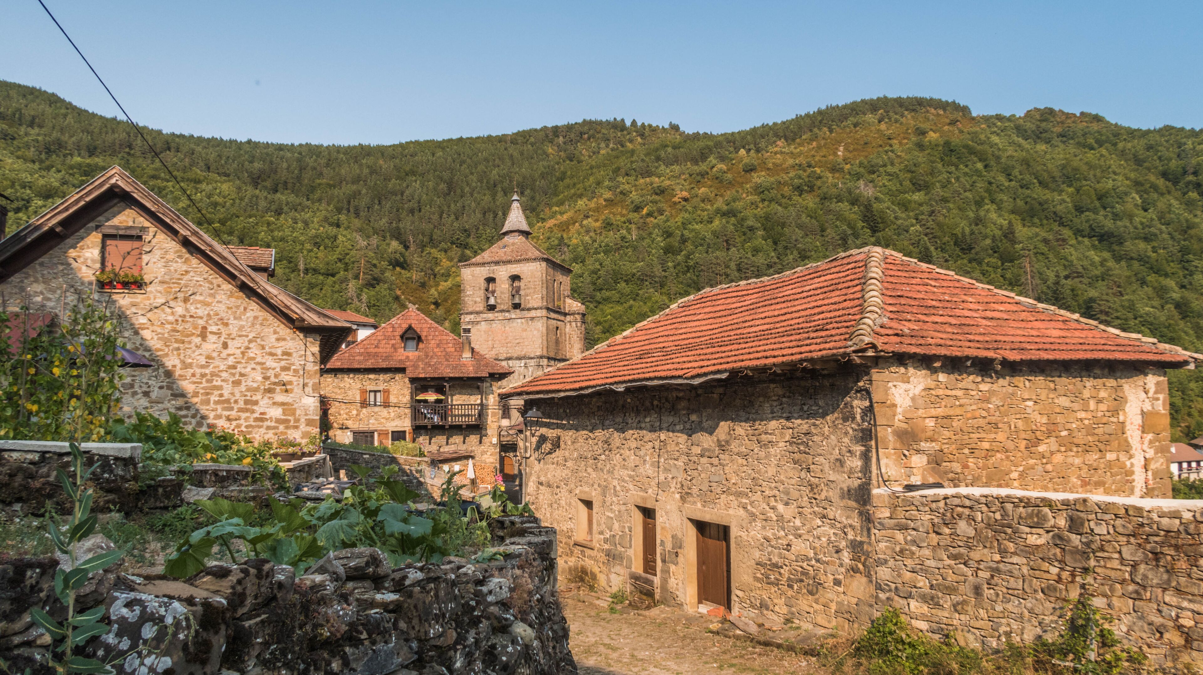 A stone building with a red-tiled roof, a bell tower, and lush green hills in the background.