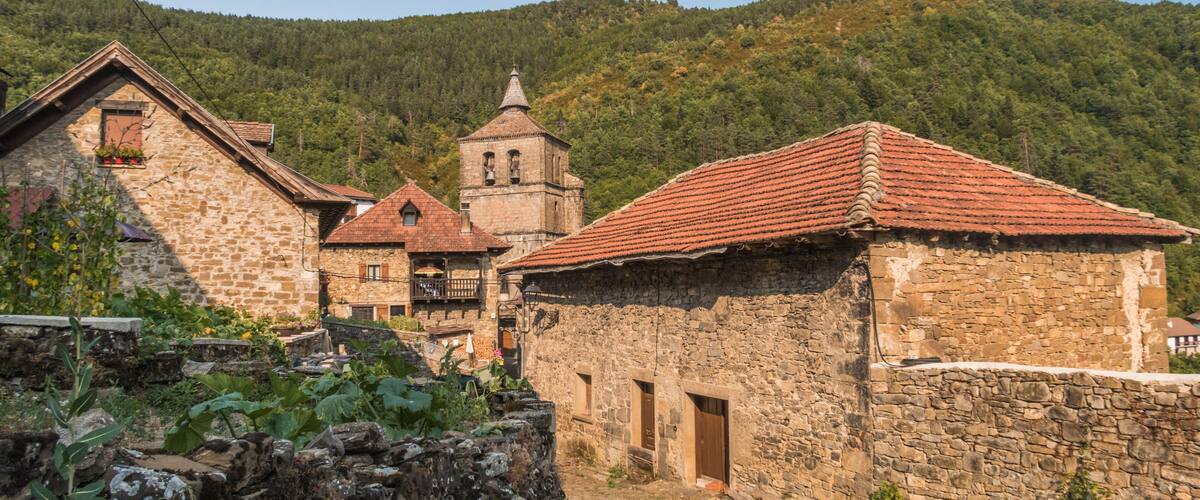 A stone building with a red-tiled roof, a bell tower, and lush green hills in the background.