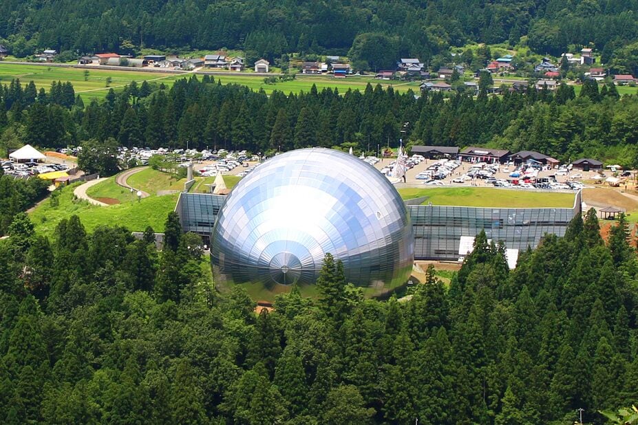 A large geodesic dome structure surrounded by a forested area and small buildings in the background.