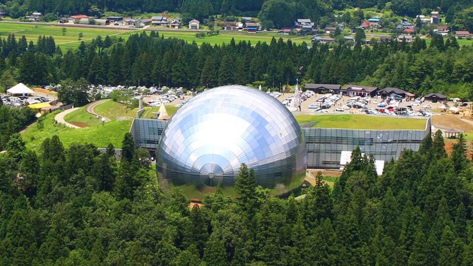 A large geodesic dome structure surrounded by a forested area and small buildings in the background.