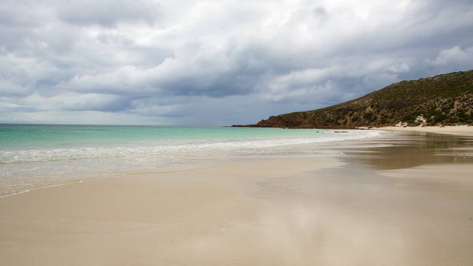 A beach with clear turquoise water, a sandy shore, and a rocky hill in the background.