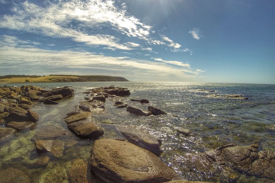 A rocky shoreline with clear water, a distant landmass, and a partly cloudy sky.