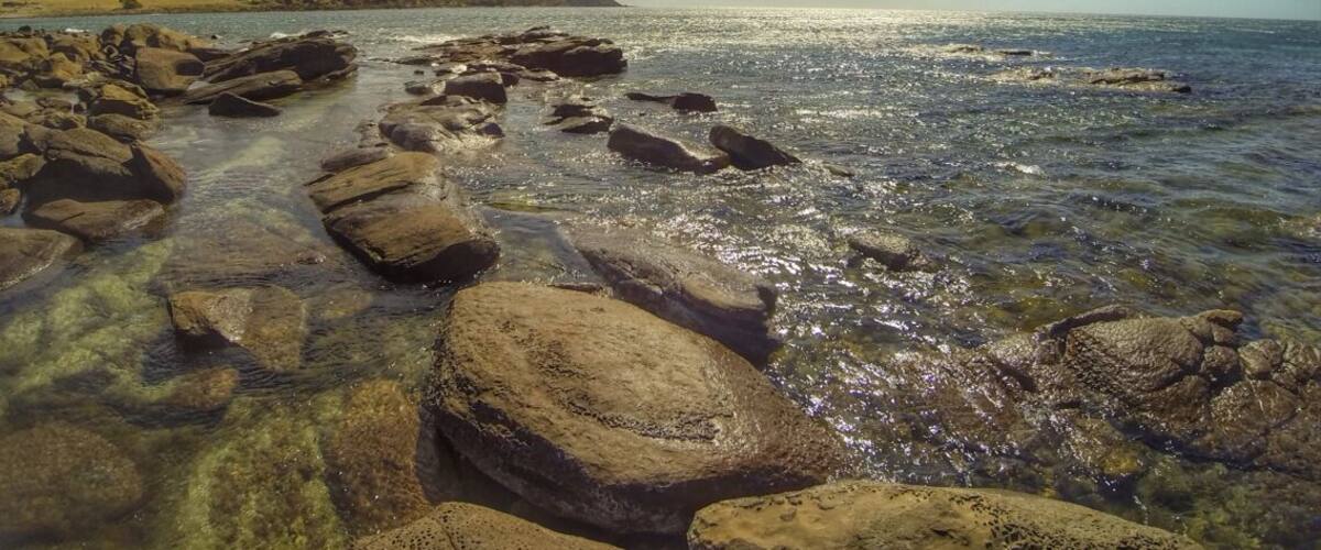 A rocky shoreline with clear water, a distant landmass, and a partly cloudy sky.