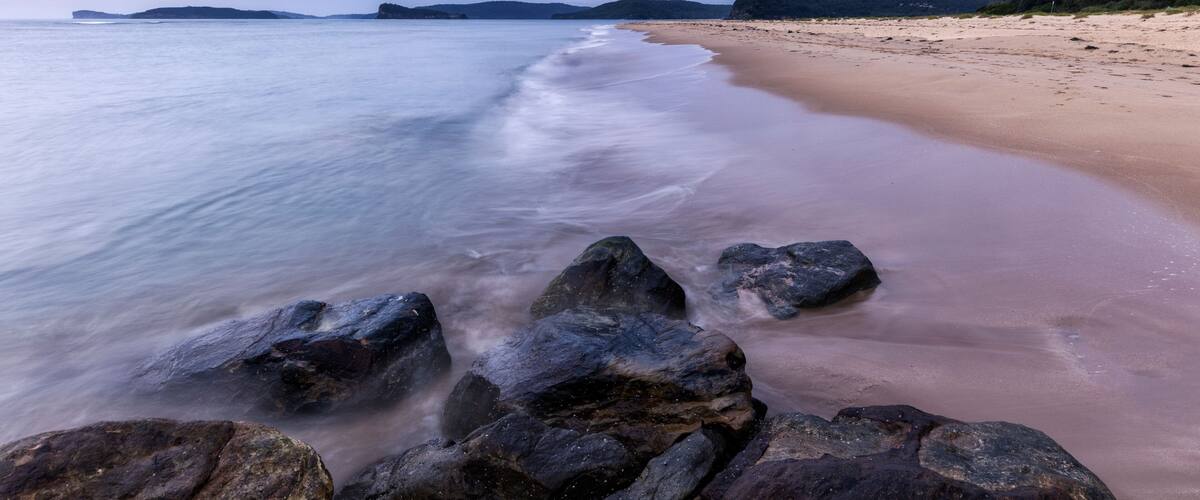 A beach with a long stretch of sand, large rocks in the foreground, and a calm sea under a cloudy sky.