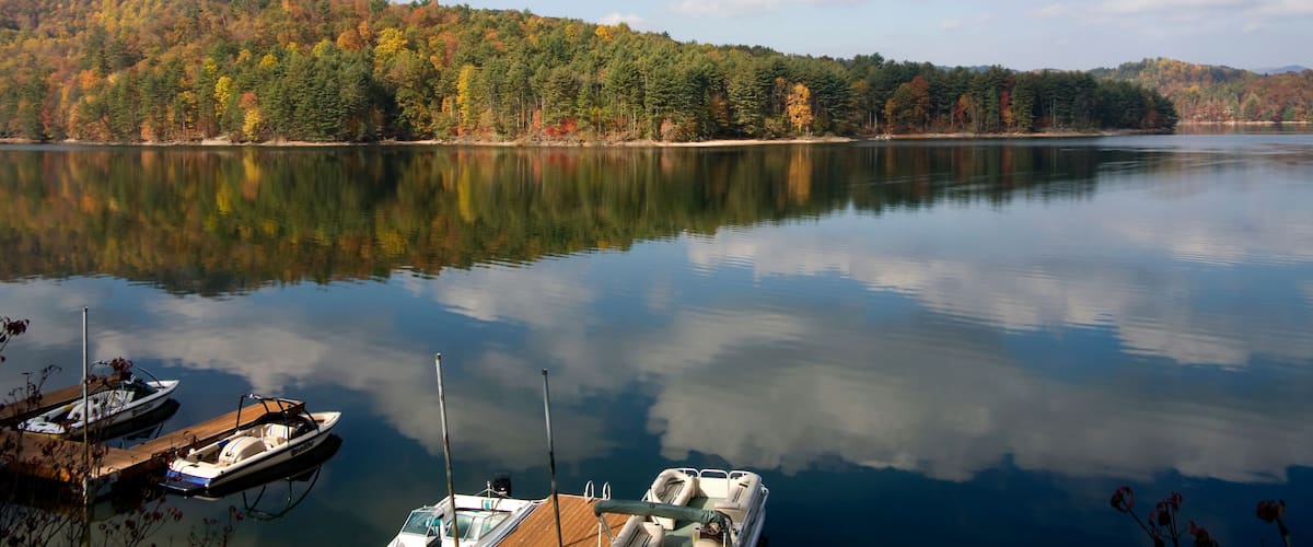 A calm lake with boats docked on the shore, surrounded by autumn-colored trees and a clear sky.