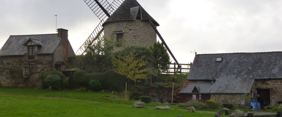 A traditional stone windmill with a wooden structure, surrounded by stone buildings and a grassy field.