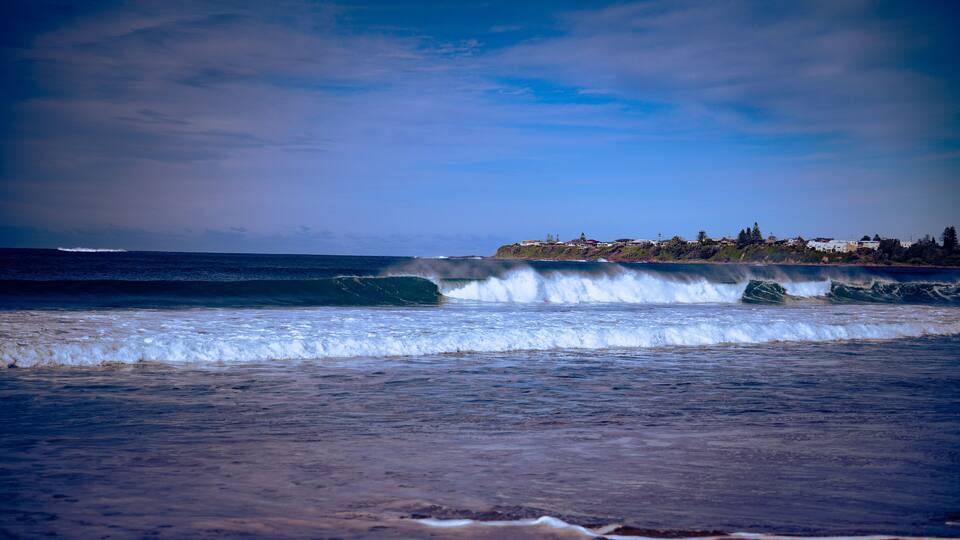A coastal landscape with waves, a clear sky, and a distant shoreline with buildings.