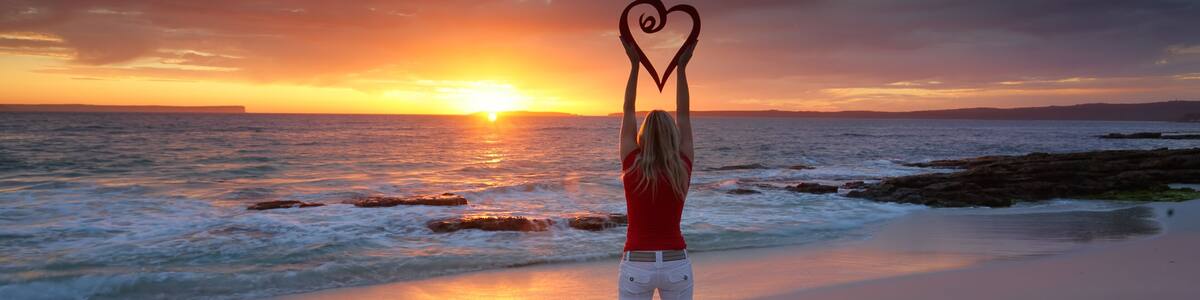 A person making a heart shape with their hands on a beach at sunset.