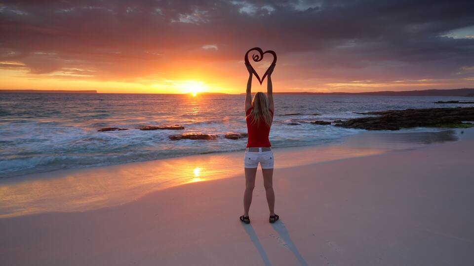 A person making a heart shape with their hands on a beach at sunset.