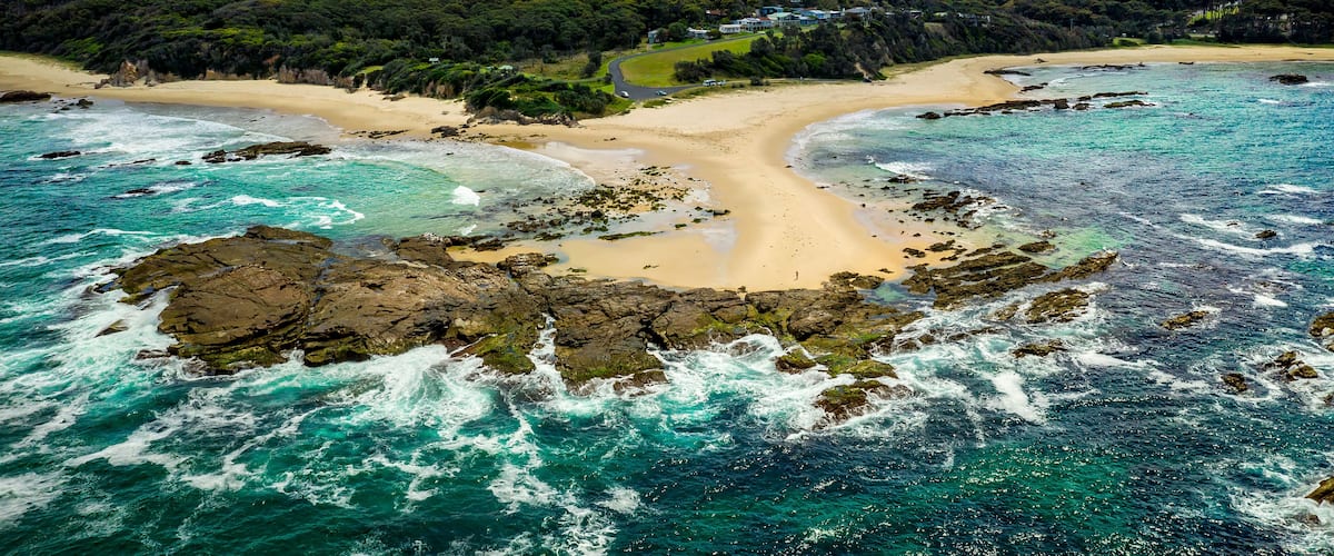 A coastal landscape with a sandy beach, rocky shoreline, turquoise waters, and a mountain in the background.