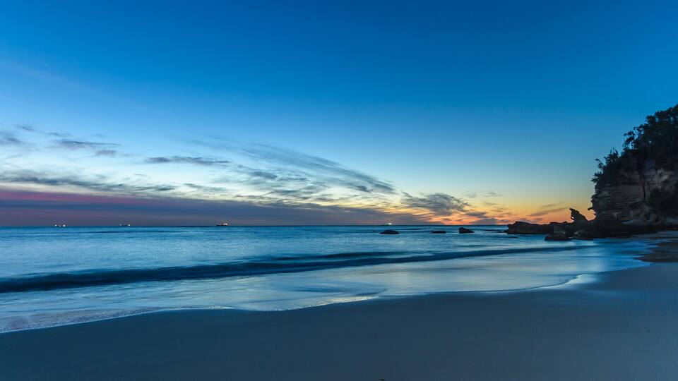 A beach with calm waves, a clear sky, and rocky cliffs.