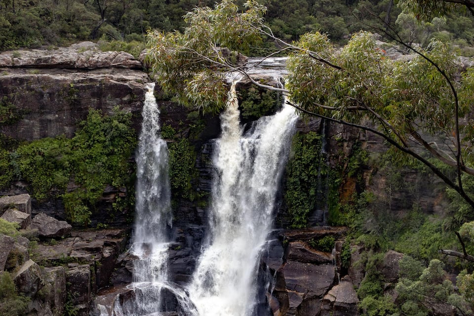 Een waterval stort neer over een rotswand, omgeven door weelderige groene bomen.