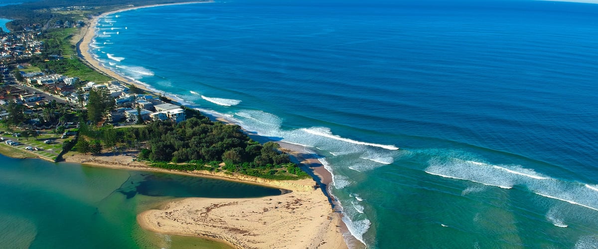 Eine Luftaufnahme einer Küstenlandschaft mit Strand, klarem blauen Meer und einer Wohngegend mit Häusern und Grünflächen.