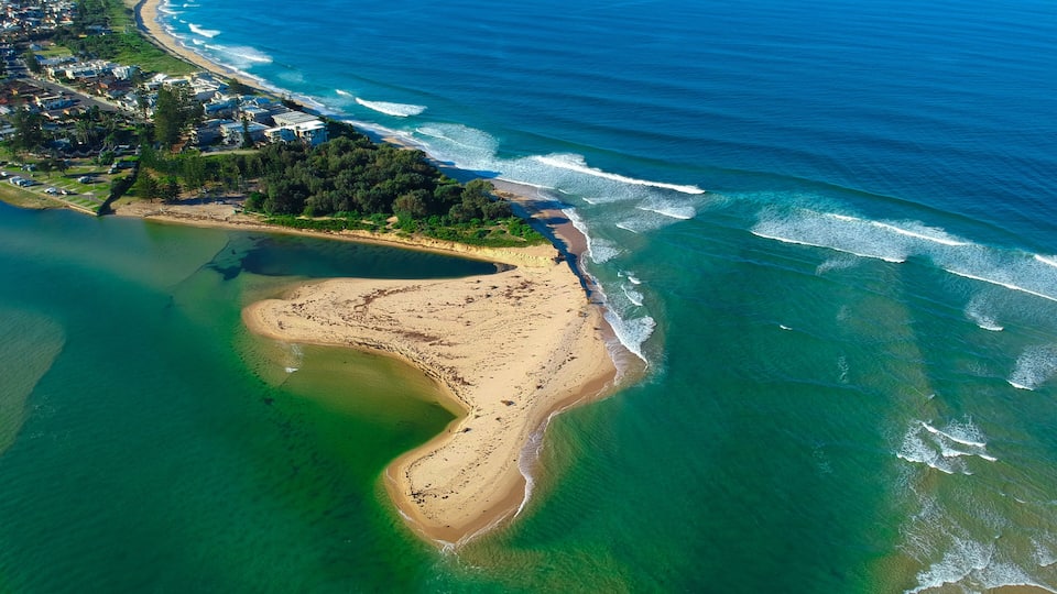 An aerial view of a coastal area with a beach, clear blue ocean, and a residential area with houses and greenery.
