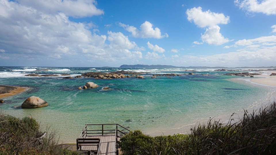 A coastal landscape with clear turquoise waters, rocky outcrops, and a wooden walkway leading to a sandy beach.
