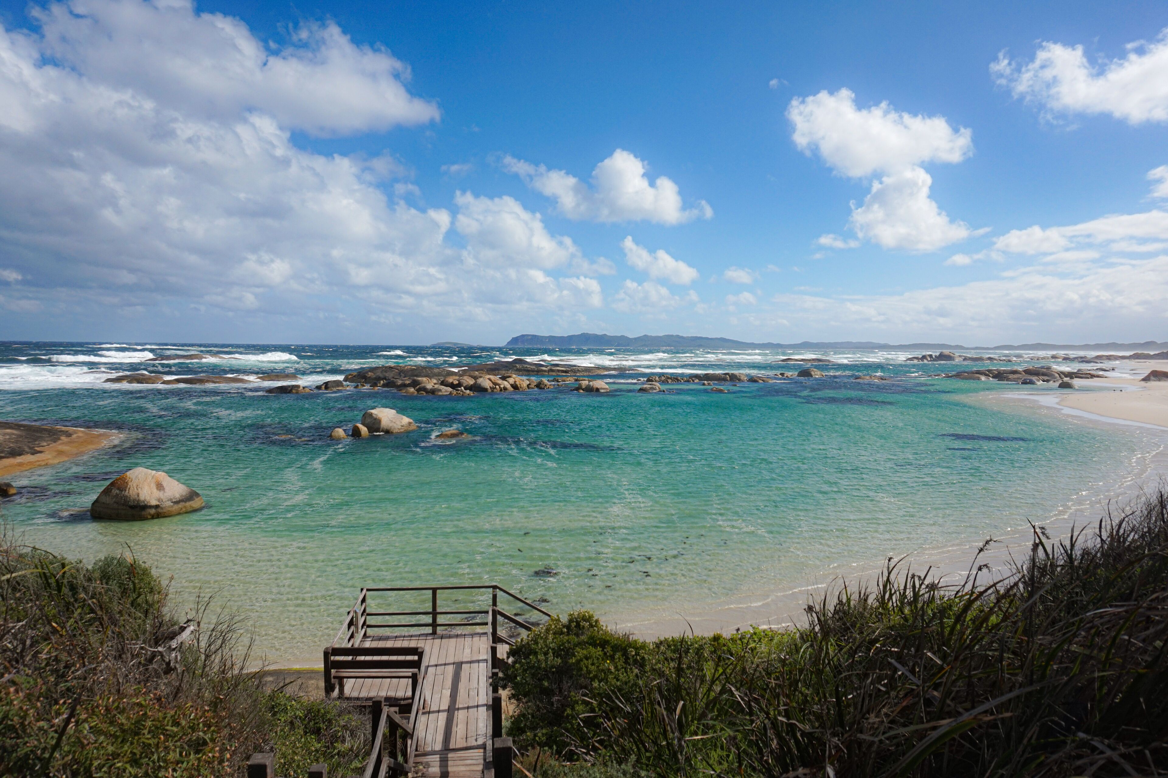 A coastal landscape with clear turquoise waters, rocky outcrops, and a wooden walkway leading to a sandy beach.